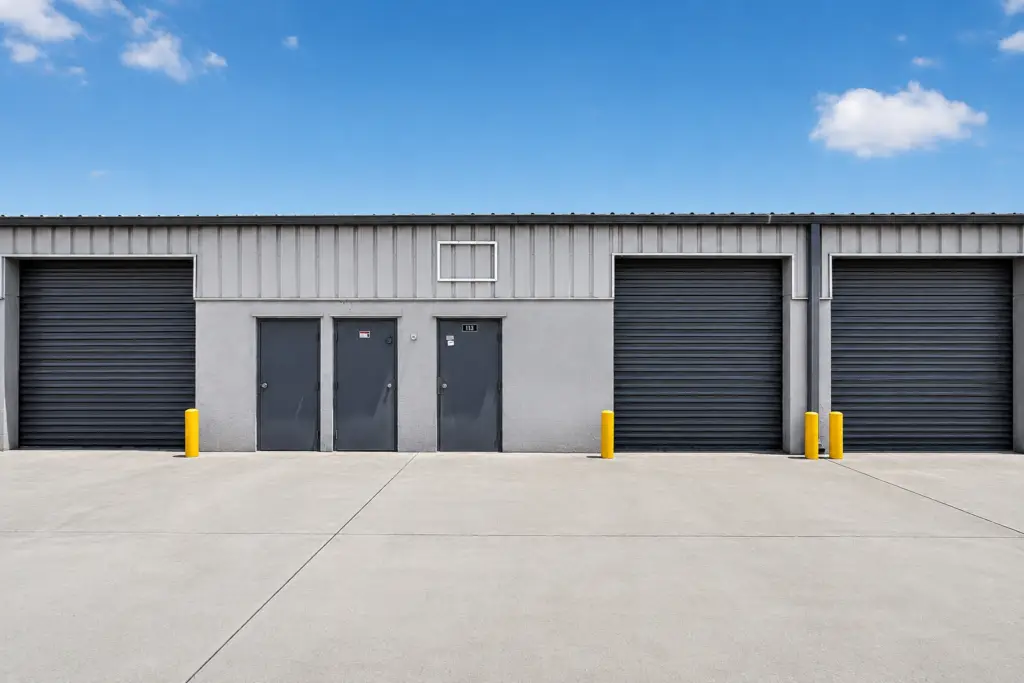 Exterior view of an industrial warehouse building with multiple roll-up doors, entry doors, and a clean concrete parking area under a blue sky.