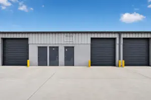 Exterior view of an industrial warehouse building with multiple roll-up doors, entry doors, and a clean concrete parking area under a blue sky.